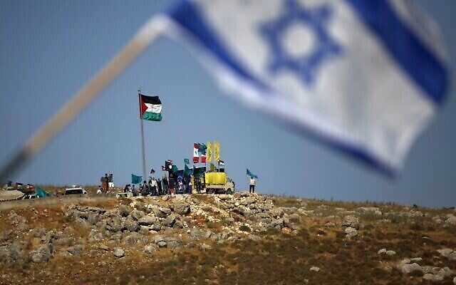 Photo prise le 20 mai 2021, depuis la ville de Metula dans le nord d'Israël, près de la frontière avec le Liban où flotte le drapeau israélien, avec en arrière-plan des gens à la périphérie du village de Kfarkila dans le sud du Liban, brandissant des drapeaux palestiniens, libanais et du Hezbollah, organisation soutenue par l'Iran. (Crédit : Jalaa Marey / AFP) Photo prise le 20 mai 2021, depuis la ville de Metula dans le nord d'Israël, près de la frontière avec le Liban où flotte le drapeau israélien, avec en arrière-plan des gens à la périphérie du village de Kfarkila dans le sud du Liban, brandissant des drapeaux palestiniens, libanais et du Hezbollah, organisation soutenue par l'Iran. (Crédit : Jalaa Marey / AFP)