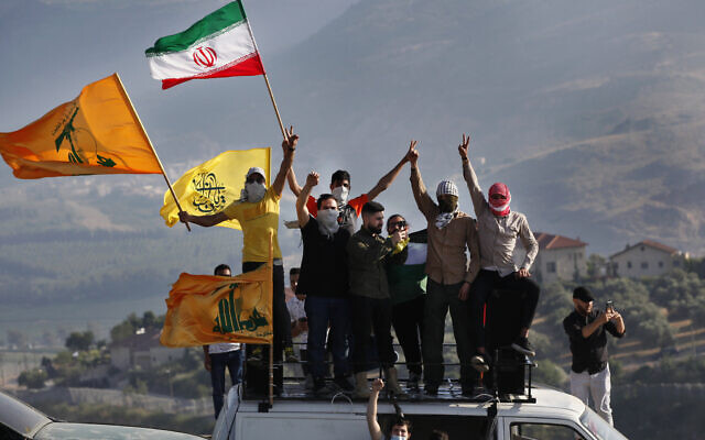 Des partisans du Hezbollah brandissent les drapeaux Palestinien, Iranien et du groupe terroriste libanais lors d'une manifestation à la frontière dans le cadre de la campagne militaire menée par les Israéliens à Gaza, à la frontière entre Israël et le Liban, devant l'implantation de Metula, en arrière-plan, le 14 mai 2021. (Crédit : AP Photo/Mohammed Zaatari)