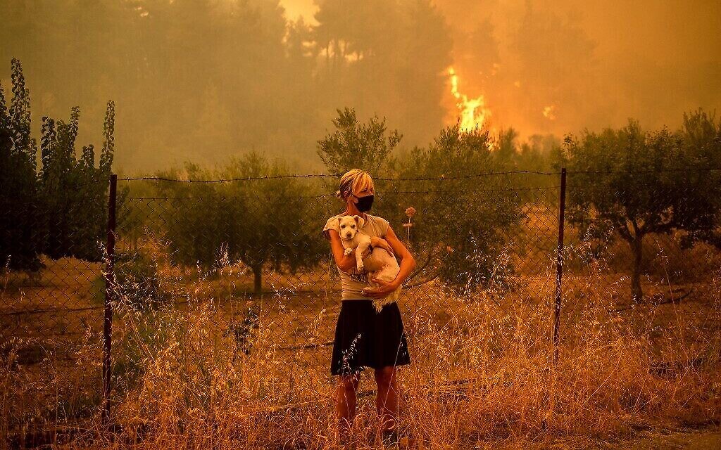 Une femme tient un chien dans ses bras alors que des feux de forêt approchent du village de Pefki sur l'île d'Evia (Eubée), la deuxième plus grande île de Grèce, le 8 août 2021. (Crédit : ANGELOS TZORTZINIS/AFP) Une femme tient un chien dans ses bras alors que des feux de forêt approchent du village de Pefki sur l'île d'Evia (Eubée), la deuxième plus grande île de Grèce, le 8 août 2021. (Crédit : ANGELOS TZORTZINIS/AFP)