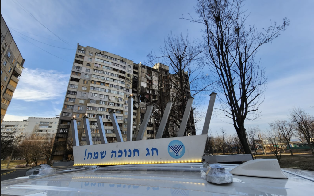  Une voiture avec une hanoukkiya fixée sur le toit passant devant un bâtiment bombardé le deuxième soir de Hanukkah, à Kharkiv, en Ukraine, le 20 décembre 2022. (Crédit : Kharkiv Choral Synagogue)