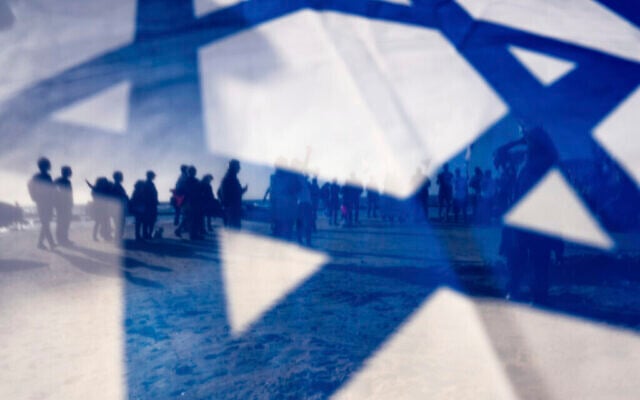 Des manifestants israéliens vus à travers le drapeau national israélien lors d’une manifestation contre le projet de réforme judiciaire du gouvernement du Premier ministre Benjamin Netanyahu, à Césarée, en Israël, le 17 mars 2023. (Crédit : AP Photo/Oded Balilty) Des manifestants israéliens vus à travers le drapeau national israélien lors d’une manifestation contre le projet de réforme judiciaire du gouvernement du Premier ministre Benjamin Netanyahu, à Césarée, en Israël, le 17 mars 2023. (Crédit : AP Photo/Oded Balilty)
