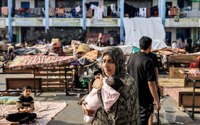 Une femme porte un nourrisson dans la cour de récréation d'une école contrôlée par l'Office de secours et de travaux des Nations unies pour les réfugiés de Palestine (UNRWA) qui a été convertie en abri pour les Palestiniens déplacés, à Khan Yunis dans le sud de la bande de Gaza, le 25 octobre 2023. (Crédit : Mahmud Hams/AFP) Une femme porte un nourrisson dans la cour de récréation d'une école contrôlée par l'Office de secours et de travaux des Nations unies pour les réfugiés de Palestine (UNRWA) qui a été convertie en abri pour les Palestiniens déplacés, à Khan Yunis dans le sud de la bande de Gaza, le 25 octobre 2023. (Crédit : Mahmud Hams/AFP)