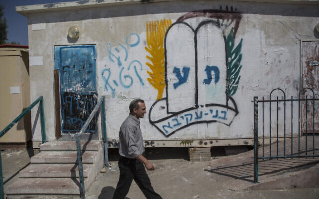 Yoram Ohev-Tzion, secrétaire de la ville de Karmei Katif, passe devant des hangars abritant le siège temporaire de la communauté Karmei Katif, près de la ville israélienne d'Amatzia, dans la région sud du mont Hébron. Sur le mur est inscrit au pochoir le logo du mouvement de jeunesse B'nei Akiva, à côté des mots « branche Katif ». 20 juillet 2015. (Crédit : Hadas Parush/Flash90) Yoram Ohev-Tzion, secrétaire de la ville de Karmei Katif, passe devant des hangars abritant le siège temporaire de la communauté Karmei Katif, près de la ville israélienne d'Amatzia, dans la région sud du mont Hébron. Sur le mur est inscrit au pochoir le logo du mouvement de jeunesse B'nei Akiva, à côté des mots « branche Katif ». 20 juillet 2015. (Crédit : Hadas Parush/Flash90)