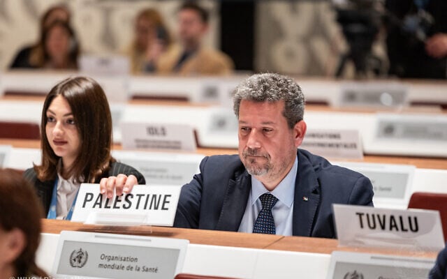 Riyad Awaja, du conseil des affaires socio-économiques à la Mission permanente d'observation de l'État de Palestine, plaçant une pancarte indiquant « Palestine » devant lui alors qu'il prend place parmi les États membres lors de la huitième séance plénière de la 77e Assemblée mondiale de la santé à Genève, en Suisse, le 1er juin 2024. (Crédit : Pierre Albouy/Organisation mondiale de la santé/AFP) Riyad Awaja, du conseil des affaires socio-économiques à la Mission permanente d'observation de l'État de Palestine, plaçant une pancarte indiquant « Palestine » devant lui alors qu'il prend place parmi les États membres lors de la huitième séance plénière de la 77e Assemblée mondiale de la santé à Genève, en Suisse, le 1er juin 2024. (Crédit : Pierre Albouy/Organisation mondiale de la santé/AFP)