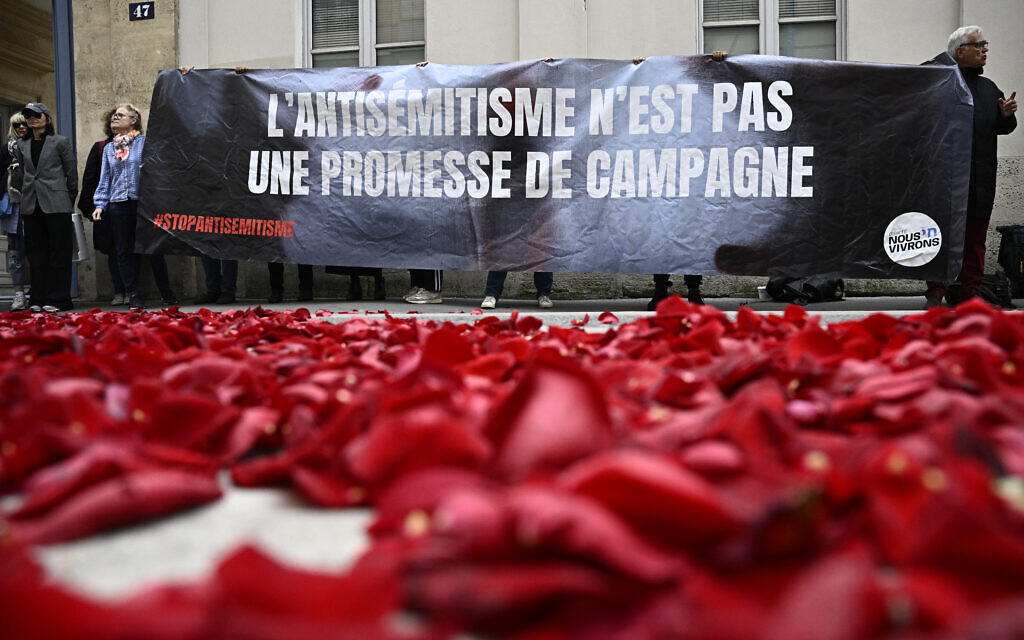 Des membres du collectif "Nous Vivrons", un groupe de lutte contre l'antisémitisme, lancent des pétales de roses pour symboliser la "fin" du parti socialiste français, alors qu'ils protestent avec une banderole "L'antisémitisme n'est pas une promesse de campagne" lors d'une manifestation devant le bâtiment de la Maison de la Chimie où le "Nouveau Front Populaire", un pacte électoral entre plusieurs partis de gauche, devrait être annoncé plus tard dans la journée, à Paris, le 14 juin 2024. (Crédit : Julien De Rosa/AFP) Des membres du collectif "Nous Vivrons", un groupe de lutte contre l'antisémitisme, lancent des pétales de roses pour symboliser la "fin" du parti socialiste français, alors qu'ils protestent avec une banderole "L'antisémitisme n'est pas une promesse de campagne" lors d'une manifestation devant le bâtiment de la Maison de la Chimie où le "Nouveau Front Populaire", un pacte électoral entre plusieurs partis de gauche, devrait être annoncé plus tard dans la journée, à Paris, le 14 juin 2024. (Crédit : Julien De Rosa/AFP)
