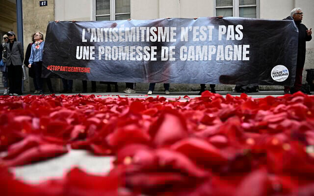 Des membres du collectif "Nous Vivrons", un groupe de lutte contre l'antisémitisme, lancent des pétales de roses pour symboliser la "fin" du parti socialiste français, alors qu'ils protestent avec une banderole "L'antisémitisme n'est pas une promesse de campagne" lors d'une manifestation devant le bâtiment de la Maison de la Chimie où le "Nouveau Front Populaire", un pacte électoral entre plusieurs partis de gauche, devrait être annoncé plus tard dans la journée, à Paris, le 14 juin 2024. (Crédit : Julien De Rosa/AFP) Des membres du collectif "Nous Vivrons", un groupe de lutte contre l'antisémitisme, lancent des pétales de roses pour symboliser la "fin" du parti socialiste français, alors qu'ils protestent avec une banderole "L'antisémitisme n'est pas une promesse de campagne" lors d'une manifestation devant le bâtiment de la Maison de la Chimie où le "Nouveau Front Populaire", un pacte électoral entre plusieurs partis de gauche, devrait être annoncé plus tard dans la journée, à Paris, le 14 juin 2024. (Crédit : Julien De Rosa/AFP)