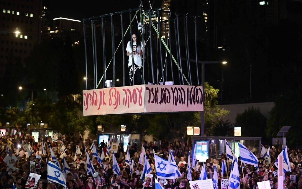 Einav Zangauker, dont le fils Matan est retenu en otage à Gaza, est suspendu à un pont dans une cage au-dessus des manifestants devant les quartiers généraux de l'armée de la Kirya, à Tel Aviv, le 7 juillet 2024. (Crédit : Tomer Neuberg/Flash90)