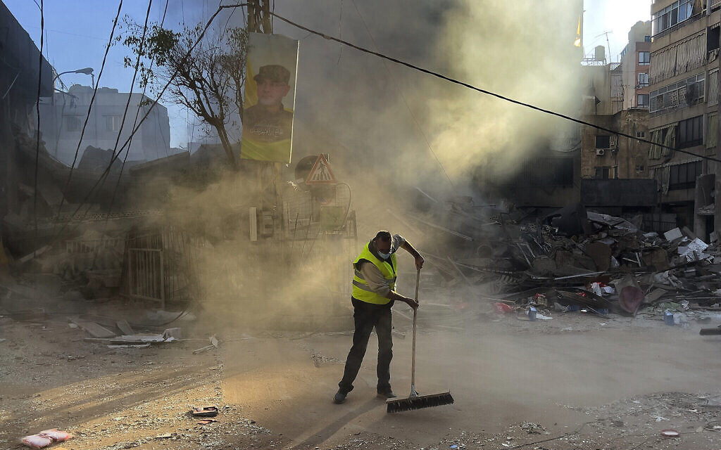 Un ouvrier nettoyant une rue alors que de la fumée s'élève d'un bâtiment détruit par une frappe aérienne israélienne à Dahiyeh, dans la banlieue sud de Beyrouth, au Liban, le 20 octobre 2024. (Crédit : Hussein Malla/AP)