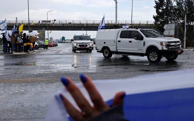 Des Israéliens brandissent le drapeau national alors qu'un convoi supposé transporter les corps des quatre otages israéliens remis par le Hamas, passe près de la ville de Sderot, dans le sud du pays, le 20 février 2025. (Crédit : John Wessels/AFP)
