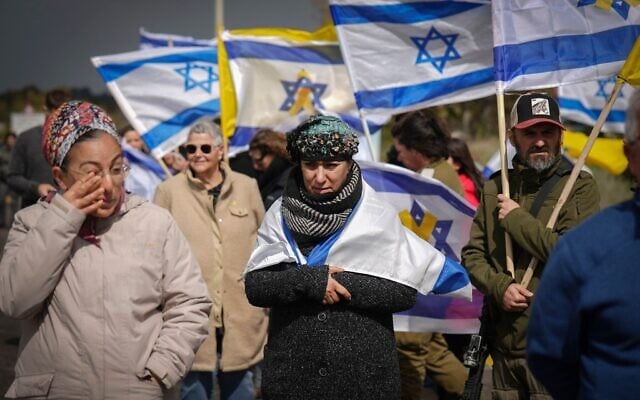 Des Israéliens brandissant des drapeaux alors qu'ils regardent passer un convoi transportant des cercueils contenant ce que l'on pense être les corps de quatre otages israéliens qui viennent d'être remis par des groupes terroristes palestiniens à Gaza, près du kibboutz Reïm, dans le sud d'Israël, le 20 février 2025. (Crédit : Ariel Schalit/AP)