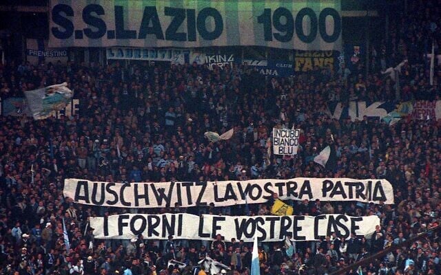 Des supporters de la Lazio déploient depuis les tribunes des banderoles sur lesquelles on peut lire : « Auschwitz est votre patrie, les fours sont vos maisons ». Les fours sont vos maisons » lors d'un match de Serie A entre la Lazio et l'AS Roma, au stade olympique de Rome, le 29 novembre 1998. 29, 1998. (Crédit : Plinio Lepri/AP) Des supporters de la Lazio déploient depuis les tribunes des banderoles sur lesquelles on peut lire : « Auschwitz est votre patrie, les fours sont vos maisons ». Les fours sont vos maisons » lors d'un match de Serie A entre la Lazio et l'AS Roma, au stade olympique de Rome, le 29 novembre 1998. 29, 1998. (Crédit : Plinio Lepri/AP)