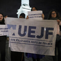 Des personnes se tiennent derrière un drapeau de l'Union des étudiants juifs de France (UEJF) lors d'un rassemblement place de la Bastille après l'agression du rabbin d'Orléans la veille, alors qu'il marchait avec son fils de neuf ans depuis la synagogue, à Paris, le 23 mars 2025. (Crédit : GEOFFROY VAN DER HASSELT / AFP)