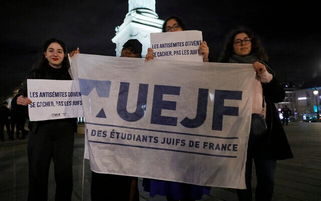 Des personnes se tiennent derrière un drapeau de l'Union des étudiants juifs de France (UEJF) lors d'un rassemblement place de la Bastille après l'agression du rabbin d'Orléans la veille, alors qu'il marchait avec son fils de neuf ans depuis la synagogue, à Paris, le 23 mars 2025. (Crédit : GEOFFROY VAN DER HASSELT / AFP) Des personnes se tiennent derrière un drapeau de l'Union des étudiants juifs de France (UEJF) lors d'un rassemblement place de la Bastille après l'agression du rabbin d'Orléans la veille, alors qu'il marchait avec son fils de neuf ans depuis la synagogue, à Paris, le 23 mars 2025. (Crédit : GEOFFROY VAN DER HASSELT / AFP)