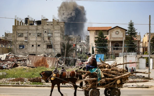 Des hommes montés sur une charrette tirée par des chevaux transportant des branches d'arbres coupées pour le bois regardent un panache de fumée s'échapper d'un bâtiment touché par une frappe israélienne après un avertissement préalable à Nuseirat, dans le centre de la bande de Gaza, le 25 mars 2025. (Crédit : Eyad BABA / AFP)