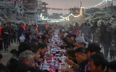 Des Palestiniens assis autour d'une grande table entourée de décombres de maisons et de bâtiments détruits pour l'iftar, le repas de rupture du jeûne, le premier jour du ramadan, à Rafah, dans le sud de la bande de Gaza, le 1ᵉʳ mars 2025. (Crédit : Abdel Kareem Hana/AP)