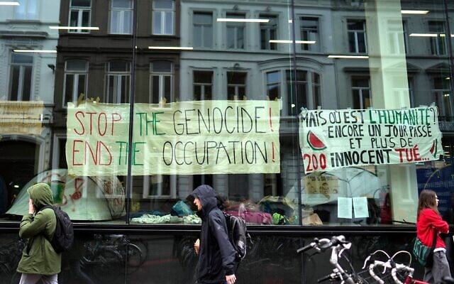 Des personnes passant devant des banderoles accrochées à l'intérieur d'une fenêtre d'un campement installé par des étudiants et des militants anti-Israël, à l'Université de Gand, alors que des étudiants occupent certaines parties du campus à Gand, en Belgique, le 16 mai 2024. (Crédit : Virginia Mayo/AP) Des personnes passant devant des banderoles accrochées à l'intérieur d'une fenêtre d'un campement installé par des étudiants et des militants anti-Israël, à l'Université de Gand, alors que des étudiants occupent certaines parties du campus à Gand, en Belgique, le 16 mai 2024. (Crédit : Virginia Mayo/AP)