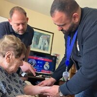 Le Dr. Fadi Badarna (à droite), ophtalmologue bénévole auprès de LeMaanam (« pour leur bien » en hébreu), une organisation à but non lucratif qui fournit des soins médicaux gratuits à domicile aux survivants de la Shoah, et Aviv Paz, employé de LeMaanam, examinant le numéro tatoué sur le bras de Chana Sandouvsky par les nazis à Auschwitz, dans sa maison de Kiryat Bialik, le 16 avril 2025. (Crédit : Diana Bletter/Times of Israel)