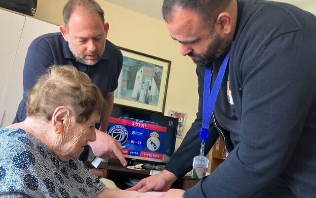 Le Dr. Fadi Badarna (à droite), ophtalmologue bénévole auprès de LeMaanam (« pour leur bien » en hébreu), une organisation à but non lucratif qui fournit des soins médicaux gratuits à domicile aux survivants de la Shoah, et Aviv Paz, employé de LeMaanam, examinant le numéro tatoué sur le bras de Chana Sandouvsky par les nazis à Auschwitz, dans sa maison de Kiryat Bialik, le 16 avril 2025. (Crédit : Diana Bletter/Times of Israel)