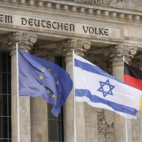 Le drapeau israélien flotte entre le drapeau européen (à gauche) et le drapeau allemand devant le bâtiment du Reichstag, siège du Bundestag, la chambre basse du Parlement allemand à Berlin, tandis que le chancelier allemande y prononce un discours sur le conflit entre Israël et le Hamas lors d'une session du Bundestag, le 12 octobre 2023. (Crédit : Odd Andersen/AFP)