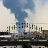 Illustration : Des drapeaux flottent le long d'un pont tandis qu'un panache de fumée et de feu s'élève d'une raffinerie de pétrole dans le sud de Téhéran, après avoir été touchée par une frappe israélienne dans la nuit du 15 juin 2025. (Crédit : ATTA KENARE / AFP)