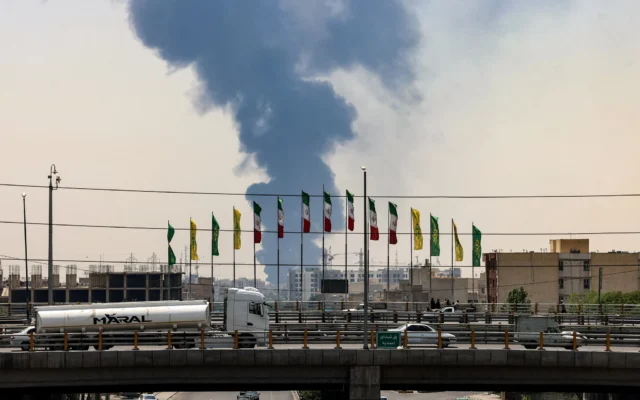 Illustration : Des drapeaux flottent le long d'un pont tandis qu'un panache de fumée et de feu s'élève d'une raffinerie de pétrole dans le sud de Téhéran, après avoir été touchée par une frappe israélienne dans la nuit du 15 juin 2025. (Crédit : ATTA KENARE / AFP)