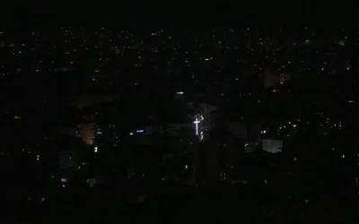 La silhouette de la croix du sommet de l'église Notre-Dame du Hadath se détache devant la banlieue de Dahiyeh à Beyrouth, en arrière-plan, plongée dans l'obscurité, au Liban, le 5 juin 2025. (Crédit : AP Photo/Hassan Ammar)