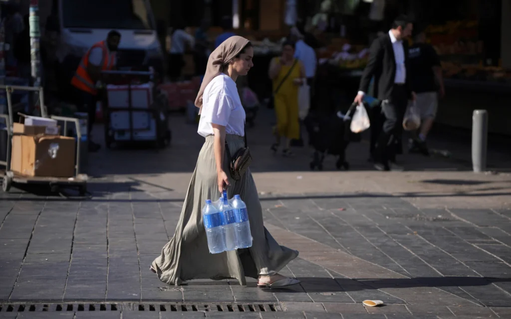 Une femme transportant de l'eau, au marché Mahane Yehuda à Jérusalem, avant le Shabbat, le 13 juin 2025. (Crédit : Ohad Zwigenberg/AP)