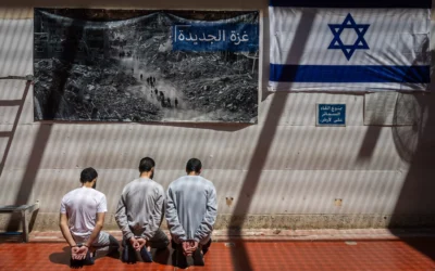 Des terroristes du Hamas menottés, assis devant une photo montrant des Palestiniens marchant parmi les décombres à Gaza et un drapeau israélien, dans une prison du centre d’Israël, le 6 mai 2025. (Crédit : Chaim Goldberg/Flash90)