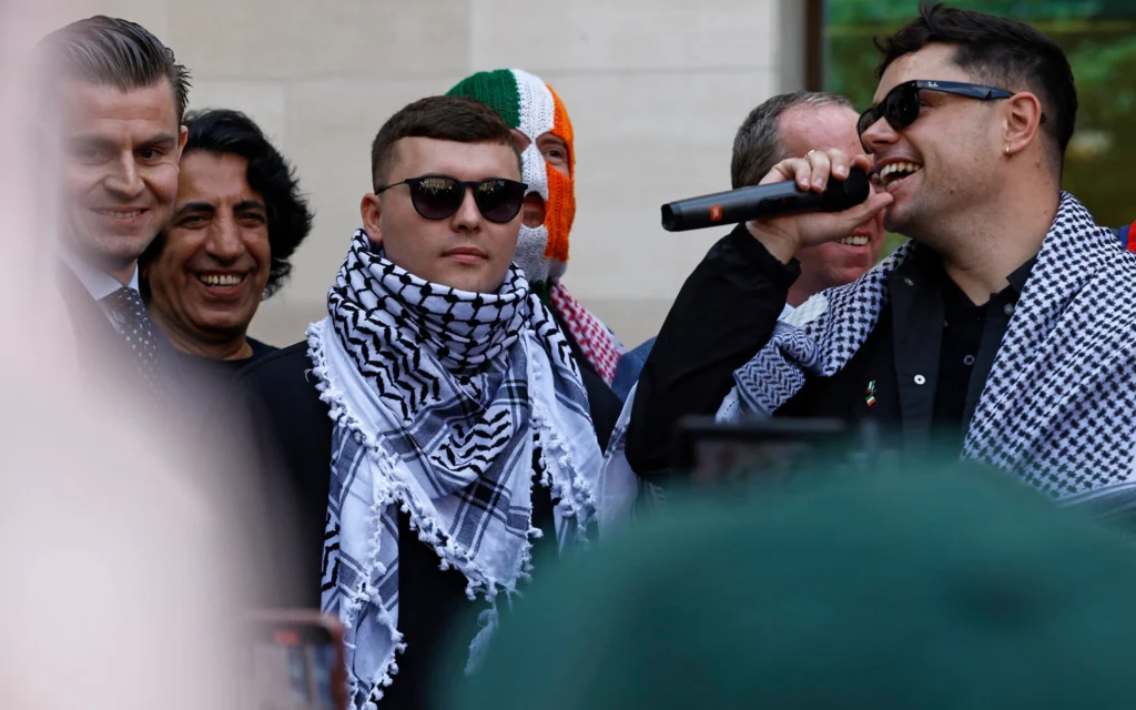 Le chanteur du groupe de rap irlandais Kneecap, Liam O'Hanna (au centre), qui se produit sous le nom de scène Mo Chara, devant le tribunal de Westminster à Londres le 18 juin 2025, après avoir été accusé de soutenir le Hezbollah. (Crédit : BENJAMIN CREMEL / AFP) Le chanteur du groupe de rap irlandais Kneecap, Liam O'Hanna (au centre), qui se produit sous le nom de scène Mo Chara, devant le tribunal de Westminster à Londres le 18 juin 2025, après avoir été accusé de soutenir le Hezbollah. (Crédit : BENJAMIN CREMEL / AFP)