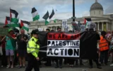 Des policiers surveillant des manifestants tenant une banderole lors d'une manifestation de soutien au groupe anti-Israël Palestine Action, à Trafalgar Square, dans le centre de Londres, le 23 juin 2025. (Crédit : Henry Nicholls/AFP)