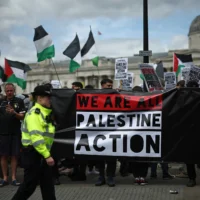 Des policiers surveillant des manifestants tenant une banderole lors d'une manifestation de soutien au groupe anti-Israël Palestine Action, à Trafalgar Square, dans le centre de Londres, le 23 juin 2025. (Crédit : Henry Nicholls/AFP)