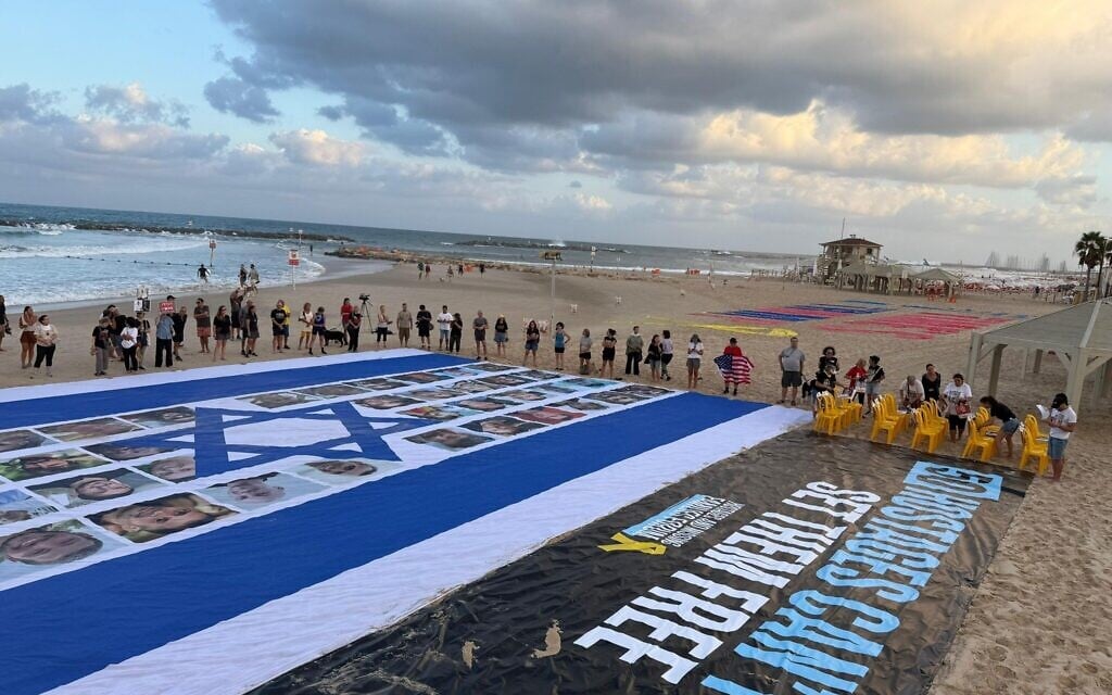 Un grand drapeau d'Israël avec les images des 50 otages restants déployé devant l'ambassade des États-Unis, à Tel Aviv, à côté d'une banderole réclamant un accord pour leur libération, le 26 août 2025. (Crédit : Aviv Atlas/Mouvement de protestation israélien pour la démocratie)