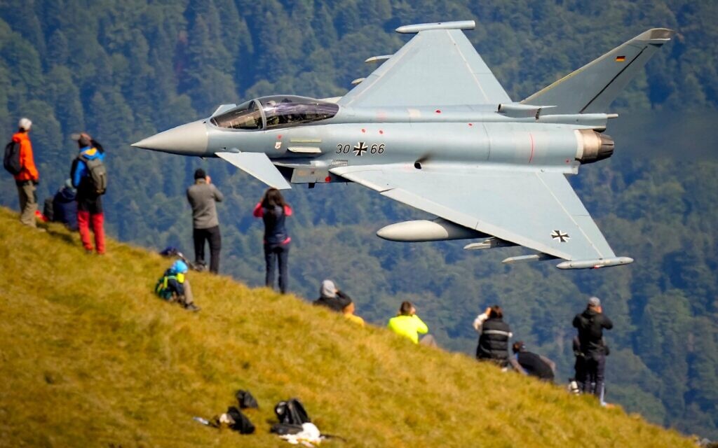 Des passionnés d'aviation observent un avion Eurofighter Typhoon de l'armée de l'air allemande passer à toute vitesse lors d'un spectacle aérien militaire et acrobatique près de la Croix des Héros, au sommet du pic Caraiman, dans les montagnes Bucegi, à Busteni, en Roumanie, le 26 août 2025. (Crédit : AP Photo/ Vadim Ghirda)