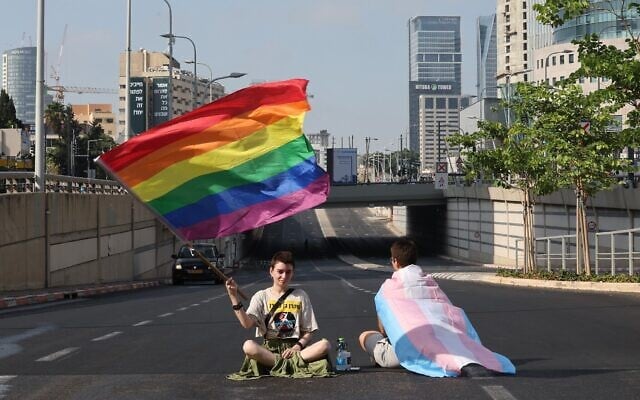 Des manifestants brandissent des drapeaux arc-en-ciel et transgenres, assis sur une route, lors d'une manifestation contre les projets de réforme judiciaire du gouvernement, à Tel Aviv, le 18 juillet 2023. (Crédit : JACK GUEZ/AFP)