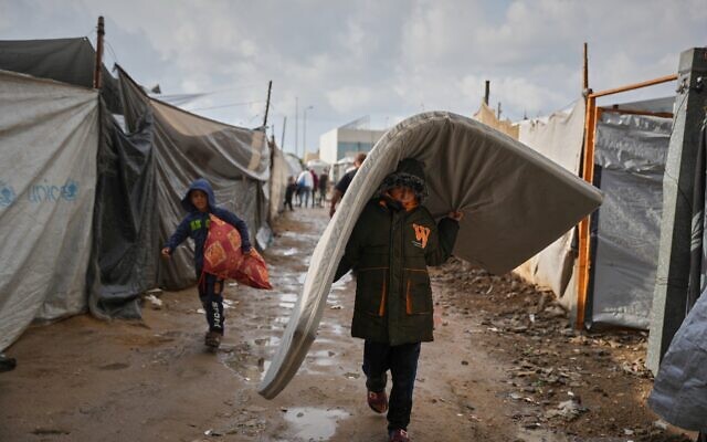 Salam Musa, 9 ans, transportant un matelas alors qu'il marche entre les tentes après la pluie dans un camp provisoire, à Deir al-Balah, dans le centre de la bande de Gaza, le 14 novembre 2025. (Crédit : Abdel Kareem Hana/AP)