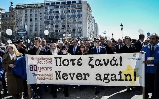Des participants, dont la présidente grecque Katerina Sakellaropoulou (au centre), défilent derrière une banderole portant les mots "Plus jamais ça" à Thessalonique, le 19 mars 2023, en route vers l'ancienne gare lors d ela commémoration du  départ du premier train qui avait déporté les Juifs grecs de Thessalonique vers le camp de concentration d'Auschwitz-Birkenau, le 15 mars 1943. (Crédit : AFP stringer/AFP)
