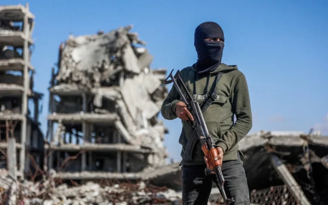 Un terroriste du Hamas montant la garde tandis que des travailleurs égyptiens, accompagnés de membres du Comité international de la Croix-Rouge (CICR), utilisent une pelleteuse pour rechercher la dépouille du dernier otage dans le camp de réfugiés de Jabaliya, dans le nord de la bande de Gaza, le 1ᵉʳ décembre 2025. (Crédit : Omar Al-Qattaa/AFP)