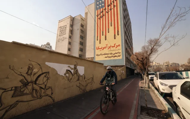 Un homme passe à vélo devant une fresque anti-américaine dans une rue de la capitale iranienne, Téhéran, le 6 janvier 2026. (Crédit : ATTA KENARE / AFP)
