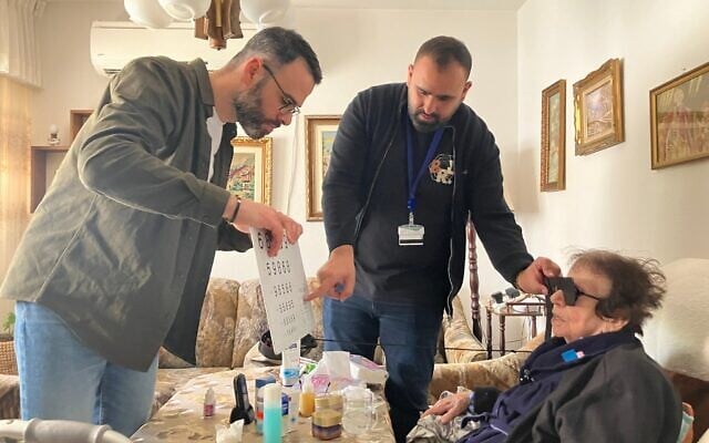 Le Dr Yosif Boulos, à gauche, et le Dr Fadi Badarna, ophtalmologues bénévoles auprès de LeMaanam ("pour leur bien" en hébreu), une organisation à but non lucratif qui fournit gratuitement des soins de santé en temps opportun aux survivants de la Shoah à leur domicile, examinent les yeux de Henia Hirsh chez elle à Kiryat Yam, le 16 avril 2025. (Crédit : Diana Bletter/Times of Israel)