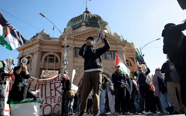 Un manifestant s'exprime à travers un mégaphone alors qu'il se tient avec d'autres personnes brandissant des drapeaux et des banderoles devant la gare de Flinders Street lors de la visite du président israélien Isaac Herzog à Melbourne, le 12 février 2026. (Crédit : MARTIN KEEP / AFP) Un manifestant s'exprime à travers un mégaphone alors qu'il se tient avec d'autres personnes brandissant des drapeaux et des banderoles devant la gare de Flinders Street lors de la visite du président israélien Isaac Herzog à Melbourne, le 12 février 2026. (Crédit : MARTIN KEEP / AFP)