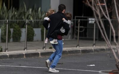 Illustration : Un homme portant deux enfants dans ses bras alors qu'il court se mettre à l'abri après le déclenchement des sirènes à Tel Aviv le 28 février 2026, à la suite de l'annonce du lancement d'une frappe préventive contre l'Iran par Israël. (Crédit : Ilia Yefimovich/AFP)