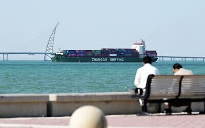 Photo d'illustration: Des personnes assises sur un banc sur la chaussée Sheikh Jaber al-Ahmad al-Sabah tandis qu'un porte-conteneurs de la compagnie Zhonggu Shipping passe devant elles en direction du port de Shuwaikh, à Koweït City, le 28 février 2026. (Crédit : Yasser al-Zayyat/AFP)