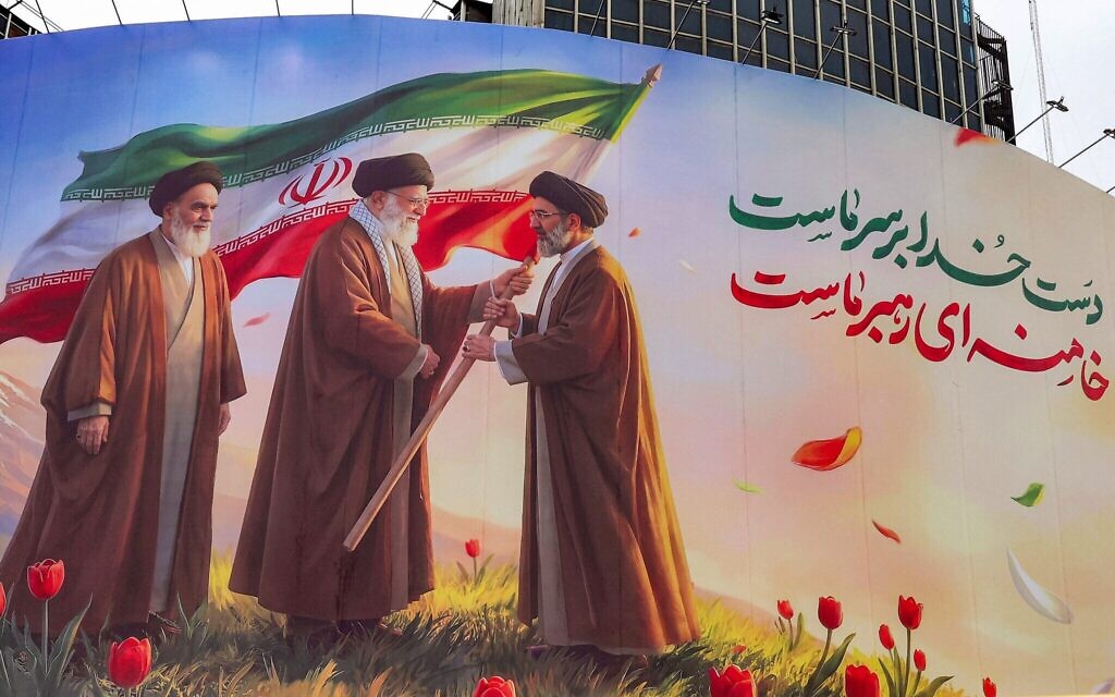 Une bannière qui représente le défunt guide suprême iranien, l'ayatollah Ruhollah Khomeini (à gauche), observant son successeur, le défunt ayatollah Ali Khamenei (au centre), remettre un drapeau de la République islamique d'Iran à son fils et nouveau guide suprême, Mojtaba Khamenei (à droite) déployée sur la place Valiasr, dans le centre de Téhéran, le 10 mars 2026. (Crédit : AFP)