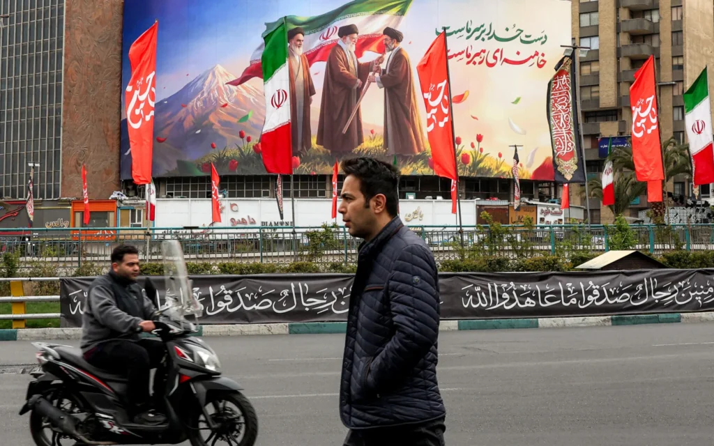 Un homme passe devant un motard près d’une banderole – représentant le défunt guide suprême iranien, l’ayatollah Ruhollah Khomeini (à gauche), observant son successeur, le défunt ayatollah Ali Khamenei (au centre), remettre un drapeau national à son fils et nouveau guide suprême, Mojtaba Khamenei (à droite) – déployée sur la place Valiasr, dans le centre de Téhéran, le 10 mars 2026. (Crédit : AFP)