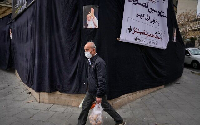 Un homme passant devant une photo du défunt Ali Khamenei, accrochée sur une bâche noire à côté d'une mosquée, les provisions à la main, à Téhéran, en Iran, le 2 mars 2026. (Crédit : Vahid Salemi/AP Photo)