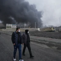 Quatre hommes marchant alors qu’un épais panache de fumée provenant d’une frappe américano-israélienne sur une installation de stockage de pétrole samedi soir persiste dans le ciel nuageux derrière eux à Téhéran, en Iran, le 8 mars 2026. (Crédit : Vahid Salemi/AP)