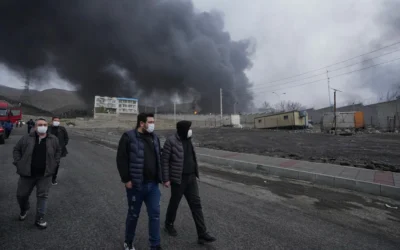 Quatre hommes marchant alors qu’un épais panache de fumée provenant d’une frappe américano-israélienne sur une installation de stockage de pétrole samedi soir persiste dans le ciel nuageux derrière eux à Téhéran, en Iran, le 8 mars 2026. (Crédit : Vahid Salemi/AP)