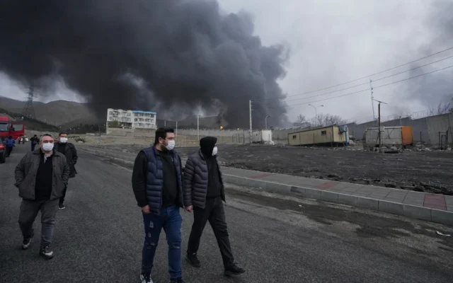 Quatre hommes marchant alors qu’un épais panache de fumée provenant d’une frappe américano-israélienne sur une installation de stockage de pétrole samedi soir persiste dans le ciel nuageux derrière eux à Téhéran, en Iran, le 8 mars 2026. (Crédit : Vahid Salemi/AP)
