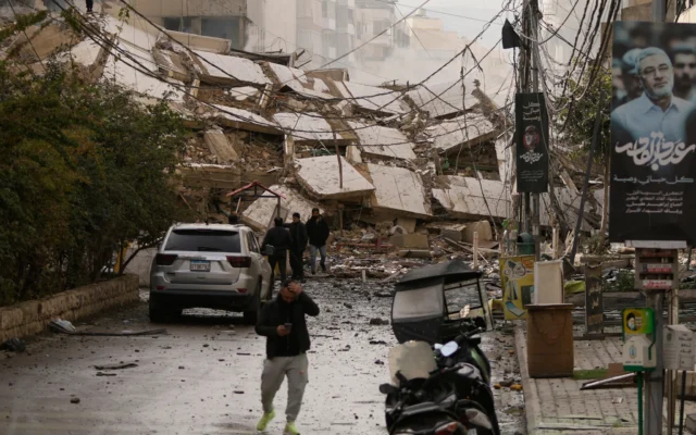Des personnes marchant dans une rue bloquée par les décombres d'un immeuble détruit lors d'une frappe aérienne israélienne, à Dahiyeh, dans la banlieue sud de Beyrouth, au Liban, le 14 mars 2026. (Crédit : Hassan Ammar/AP Photo)