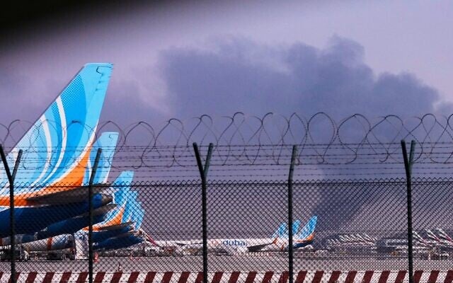 Des avions de FlyDubai stationnés à l'aéroport international de Dubaï tandis que de la fumée s'élève en arrière-plan après que le drone iranien a frappé un réservoir de carburant, entraînant la suspension temporaire des vols, à Dubaï, aux Émirats arabes unis, le 16 mars 2026. (Crédit : AP Photo)